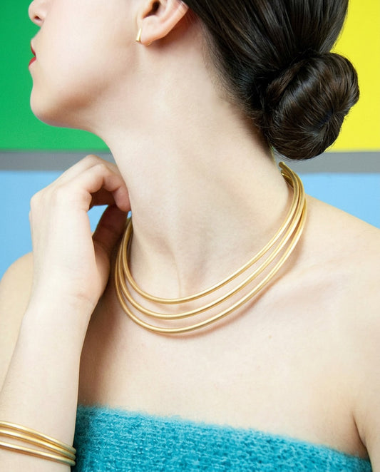 Gargantilla Sumatra triple dorada mate en el cuello de una modelo de perfil, con jersey turquesa y fondo de colores