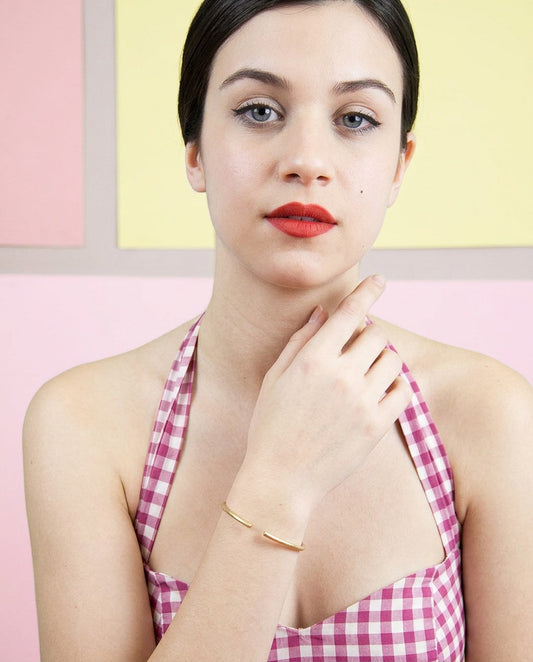 Modelo con la pulsera Sumatra sencilla dorada en la muñeca y la mano en el cuello, top de cuadros rosas